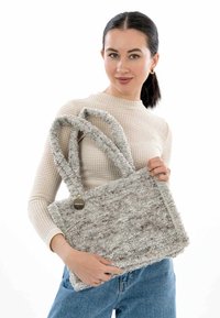 Borsa tote grigia testurizzata con due manici in peluche e un'etichetta metallica rotonda, tenuta da una donna in un maglione chiaro e jeans blu.