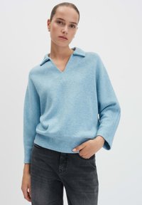 Vrouw in een lichtblauwe V-halstrui met kraag en donkergrijze spijkerbroek, staand met één hand in haar zak tegen een effen achtergrond.