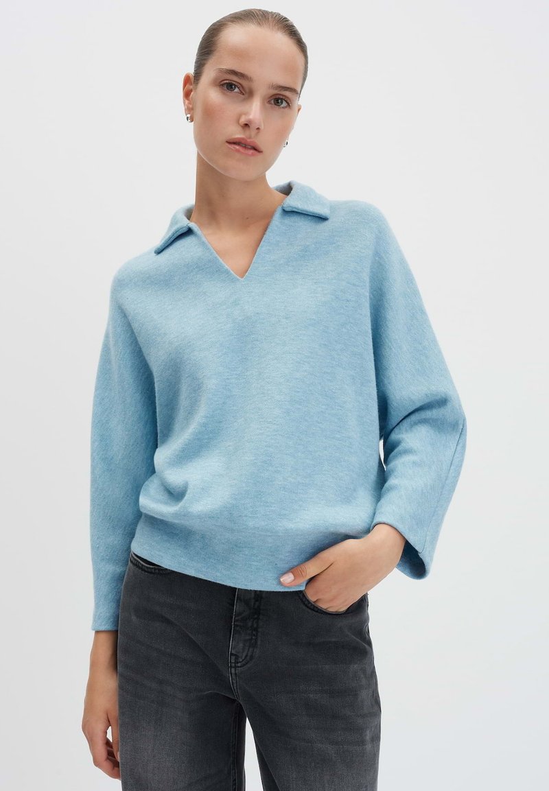 Vrouw in een lichtblauwe V-halstrui met kraag en donkergrijze spijkerbroek, staand met één hand in haar zak tegen een effen achtergrond.