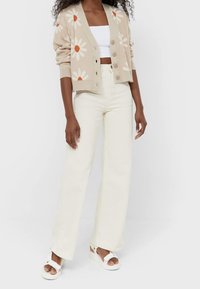 Kobieta w beżowym kardiganie z białymi stokrotkami, białym topie crop, wysokich kremowych spodniach i białych sandałach, stojąca na jednolitym tle.