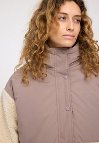 Padded jacket in muted taupe with a faux shearling beige sleeve; features a high collar and button closure. "MAZINE" logo on chest.