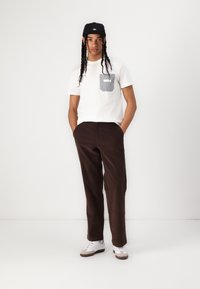 Tricou alb cu un buzunar în dungi, pantaloni largi maro și sneakers albi cu accente gri. Modelul poartă o șapcă neagră și stă într-o poziție relaxată.