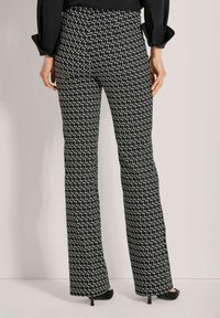 Vrouw in zwart-witte geometrisch gepatterneerde broek, zwarte hoge hakken en een zwart lange mouwen top, staande tegen een effen achtergrond.