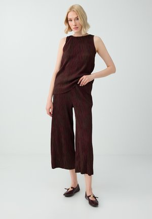 Femme aux cheveux blonds courts portant un haut sans manches marron foncé texturé et un pantalon large court avec des chaussures foncées sur fond blanc.