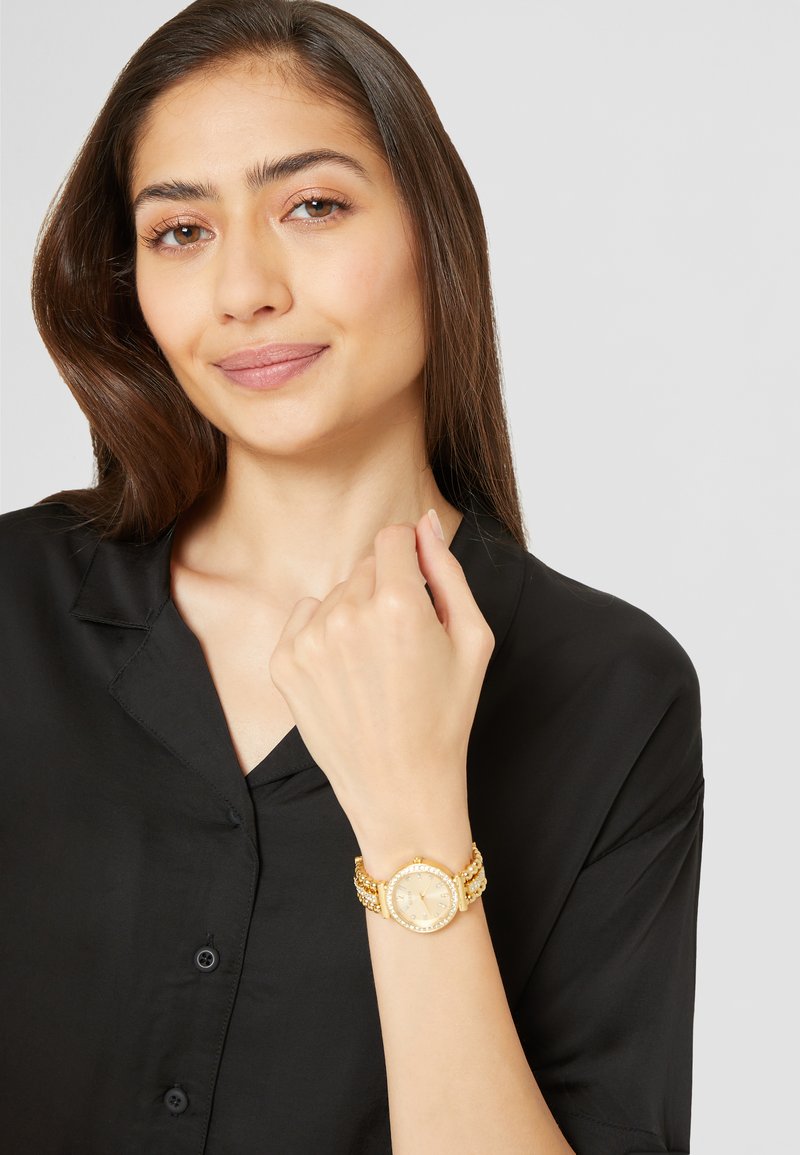 Gold-tone watch with a round case and a textured band featuring pearl accents, worn on a wrist with a black button-up shirt.