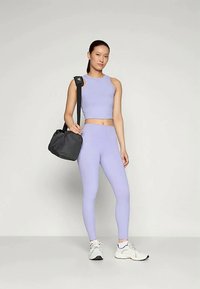 Világoslila sportos szett, amely egy ujjatlan crop topból és magasan hordott leggingsből áll. Fekete sporttáska és fehér sportcipők. Sima textúra.