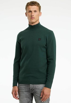 Dunkelgrüner Rollkragenpullover aus weichem, strukturiertem Strick. Enthält ein kleines schwarzes Logo-Patch auf der Brust und eng anliegende Ärmel. Wird mit grauen Jeans kombiniert.