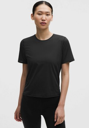 Młoda kobieta z czarnymi włosami związanymi do tyłu, ubrana w prostą czarną koszulkę z krótkim rękawem i czarne spodnie, stojąca na białym tle.