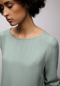 Haut vert clair à manches longues avec un col rond et une texture lisse, présentant un drapé doux aux épaules et des détails de couture minimaux.