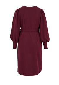 Bordeauxrode jurk van gladde stof, met lange pofmouwen, ronde halslijn, ceintuur in de taille en een lengte tot aan de knie. Geen zichtbare patronen of accessoires.