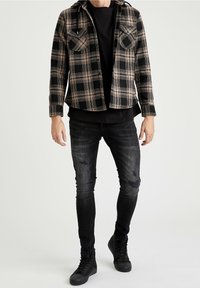 Rutig flanneljakke i sort og tan med to brystlommer; båret over en sort T-shirt. Slim-fit, slidte sorte jeans og sorte sneakers.