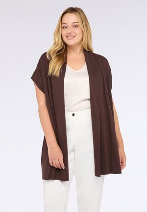 Femme souriante avec de longs cheveux blonds portant un cardigan brun ouvert sur une blouse blanche et un pantalon blanc, sur un fond uni.