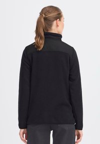 Schwarze Fleecejacke mit hohem Kragen, ausgestattet mit einem glatten, schwarzen Paneel über dem oberen Rücken. Lange Ärmel mit lockerer Passform.