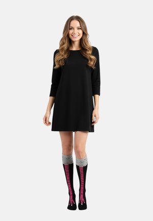 Femme souriante portant une robe noire et des chaussettes montantes conçues pour ressembler à des pattes d'oiseau avec des pieds roses et des dessus gris.