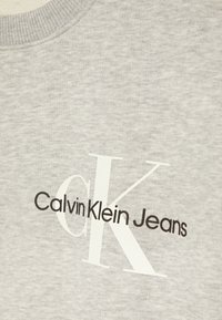 Sudadera gris con acabado texturizado, con una gran "K" blanca y el logotipo "Calvin Klein Jeans" en negro. Diseño de cuello redondo.