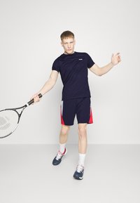 Camiseta atlética de color navy con mangas cortas y un logo sutil; combinada con pantalones cortos navy y rojos. Sosteniendo una raqueta de tenis. Zapatillas deportivas.