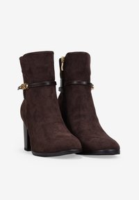 Botas de ante marrón hasta el tobillo con punta afilada, cremallera lateral y una correa contrastante con hebilla en tono dorado. Tacón alto y cuadrado.