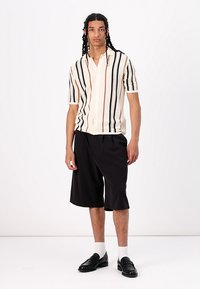 Leichte kurzärmlige Button-Up-Bluse mit abwechselnden schwarzen und beige Streifen; kombiniert mit lockeren schwarzen Shorts und schwarzen Slippern.