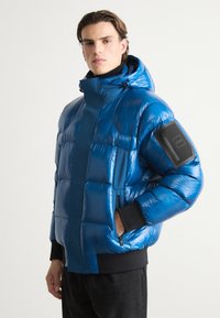 Pūdera jaka spožā elektriski zilenā krāsā ar augstu apkakli, ar rāvējslēdzēja kabatām un melniem akcentiem uz piedurknēm. Tekstūras, quilted dizains.
