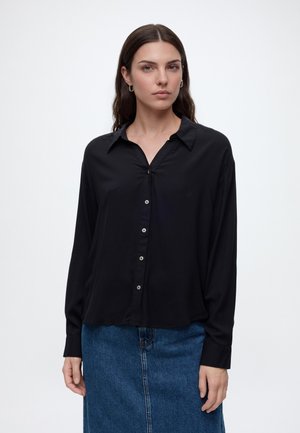 Femme aux longs cheveux bruns portant une chemise noire boutonnée et une jupe en jean bleue, debout devant un fond blanc uni.