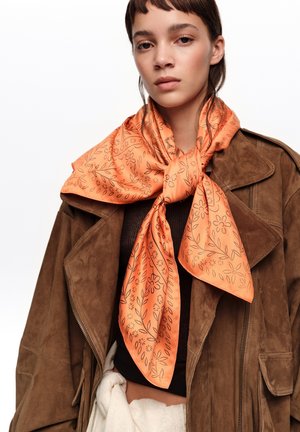 Mujer joven que lleva una chaqueta de ante marrón y un pañuelo de seda con estampado floral naranja atado alrededor del cuello, sobre un fondo blanco.