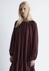Braunes, langärmliges Kleid mit gerafftem Ausschnitt und gestuftem Rüschensaum. Weicher Stoff und lockerer Schnitt erzeugen eine fließende Silhouette.