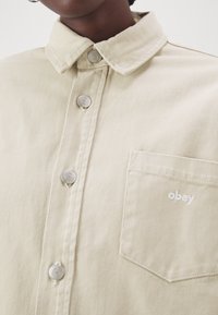 Beige denim shirt with metallic buttons and a chest pocket embroidered with the word "obey," worn by a person with dark skin.