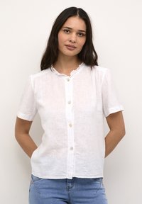 Hvid kortærmet bluse lavet af linned, med en rund, volangkrave og små knapper ned ad fronten, parret med blå jeans.