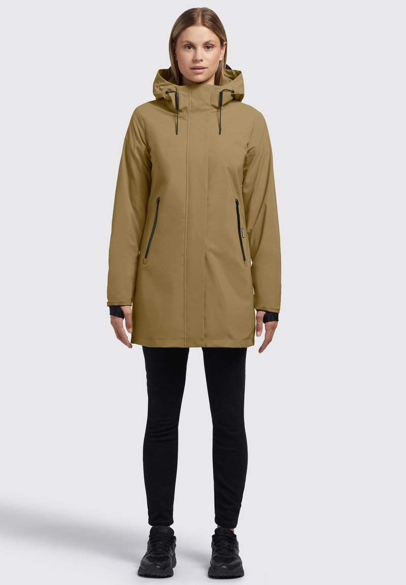 Veste à capuche kaki à coupe droite, fermeture éclair sur le devant, deux poches latérales et accents noirs. Portée par-dessus des leggings noirs et des baskets.