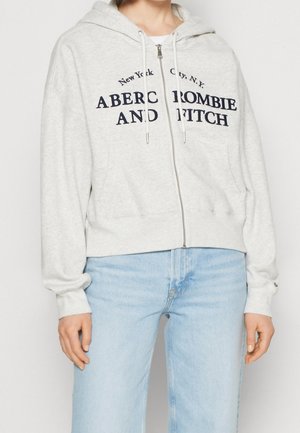 Personne portant un sweat à capuche gris clair à fermeture éclair avec "Abercrombie and Fitch" et un jean bleu clair, debout devant un fond uni.