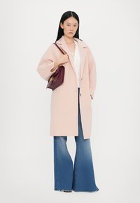 Cappotto in lana rosa chiaro con una vestibilità rilassata, maniche ampie e due bottoni neri. Abbinato a jeans a zampa d'elefante blu e una borsa bordeaux.