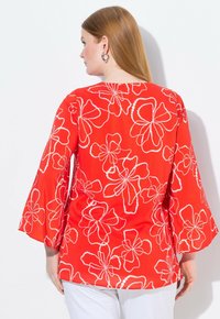 Blouse rouge avec un motif floral blanc, manches évasées et coupe décontractée, fabriquée dans un tissu léger adapté aux temps chaud.