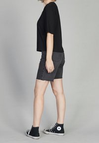 Camiseta negra corta, pantalones cortos de mezclilla grises con bordes deshilachados y zapatillas negras de caña alta. Modelo de lado contra un fondo gris.