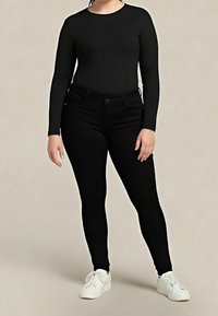 Personne debout portant un haut noir à manches longues, un pantalon noir ajusté et des baskets blanches sur un fond beige uni.