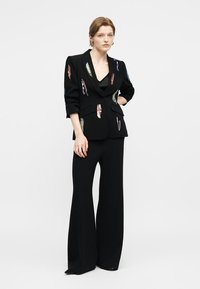 Γυναίκα που φοράει μαύρο σακάκι με πολύχρωμα παγιέτα patches σε συνδυασμό με μαύρο παντελόνι φαρδιάς γραμμής, όρθια μπροστά σε ένα απλό λευκό φόντο.