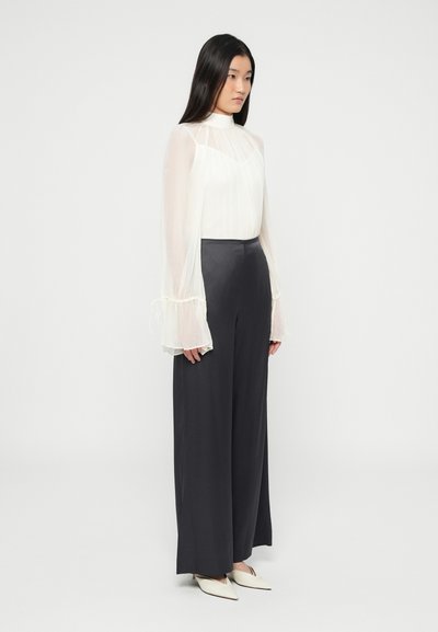 Blouse blanche transparente à col montant et manches longues évasées, associée à un pantalon large foncé. La tenue présente une texture lisse et un design minimaliste.