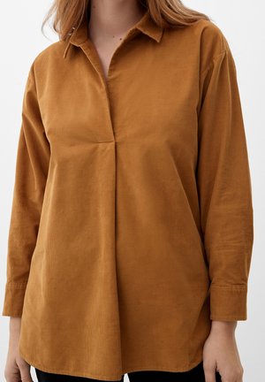 Vrouw draagt een langemouwen, loszittend, bruin corduroy overhemd met een kleine kraag tegen een eenvoudige lichte achtergrond.