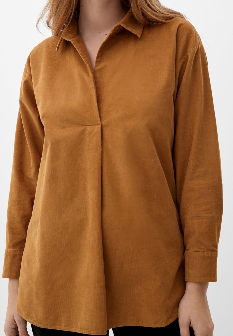 Femme portant une chemise en velours côtelé marron à manches longues, coupe ample, avec un petit col, sur un fond clair uni.