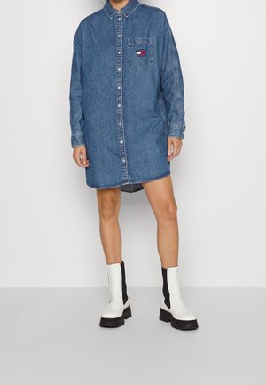 Osoba ubrana w zapinaną na guziki niebieską sukienkę dżinsową z kieszenią na piersi z logo oraz białe botki za kostkę z czarnymi elastycznymi bokami.