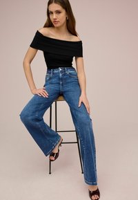 Top negru cu umeri dezgoliți, cu textură în dungi, asortat cu blugi evazați din denim albastru, cu talie înaltă, având o croială relaxată și un aspect mai deschis, completat cu sandale cu toc.