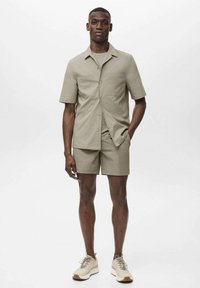 Lyse beige kortærmet skjorte med knapper og matchende shorts, lavet af glat stof. Båret med beige sneakers, der viser et minimalistisk design.