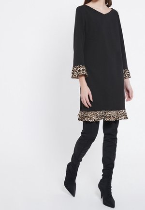 Kvinde iført en sort kjole med leopardprint flæsekant ved ærmer og kant, kombineret med sorte over-the-knee støvler med hæl.