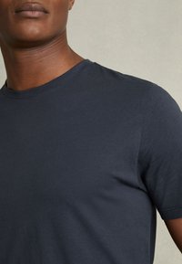 Lähikuva henkilöstä, joka pukeutunut yksiväriseen tummaan laivaston siniseen pyöreäkaulaiseen T-paitaan neutraalia taustaa vasten.