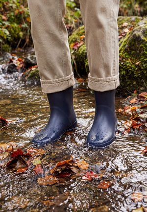 Une personne portant un pantalon beige retroussé et des bottes en caoutchouc bleu marine se tient dans un ruisseau peu profond jonché de feuilles d’automne.