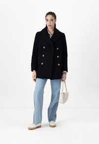 Abrigo de paño negro de doble botonadura con botones dorados, combinado con jeans de pierna ancha azul claro y zapatillas blancas; sosteniendo un pequeño bolso blanco.