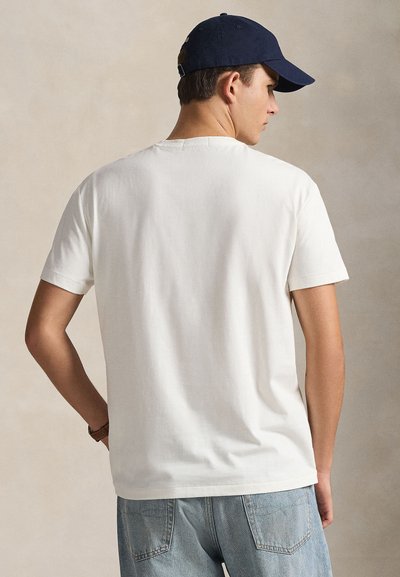 Λευκό βαμβακερό t-shirt με κοντά μανίκια και στρογγυλή λαιμόκοψη. Φορεμένο με ανοιχτόχρωμο τζιν, εμφανιζόμενο από πίσω, με ένα μπλε σκούρο καπέλο του μπέιζμπολ.