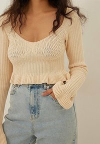 Beige pletený zkrácený svetr s lodičkovým výstřihem, texturovaným vzorem a rozšířenými rukávy, spárovaný s světle modrými džíny s vysokým pasem.