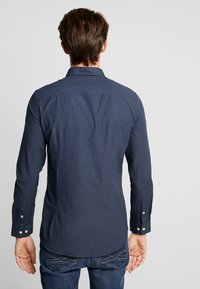 Man som vänder ryggen mot kameran och bär en mörkblå mönstrad skjorta med långa ärmar samt blå jeans mot en enkel ljus bakgrund.