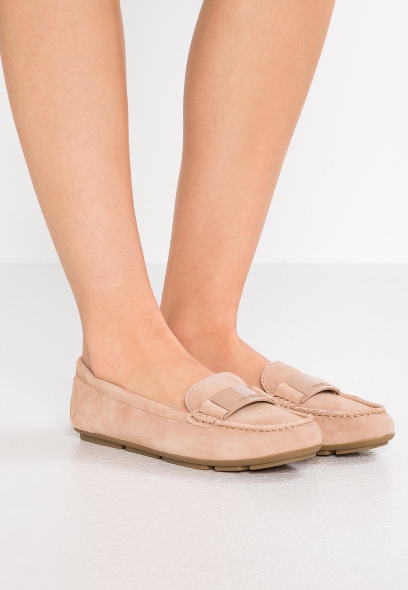 Pieds chaussés de mocassins en daim beige avec semelles en caoutchouc et un petit détail métallique de marque sur le dessus, debout sur un sol blanc.