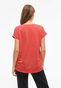 Kortærmet koralfarvet T-shirt med rullede manchetter, blød tekstur og afrundet kant, parret med sorte bukser, der viser bagsiden.